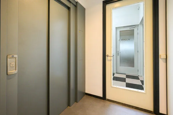 Hal met gesloten liftdeuren en een deur met raam dat uitzicht biedt op een gang met de naam 'VANDIJK' op een glazen deur.